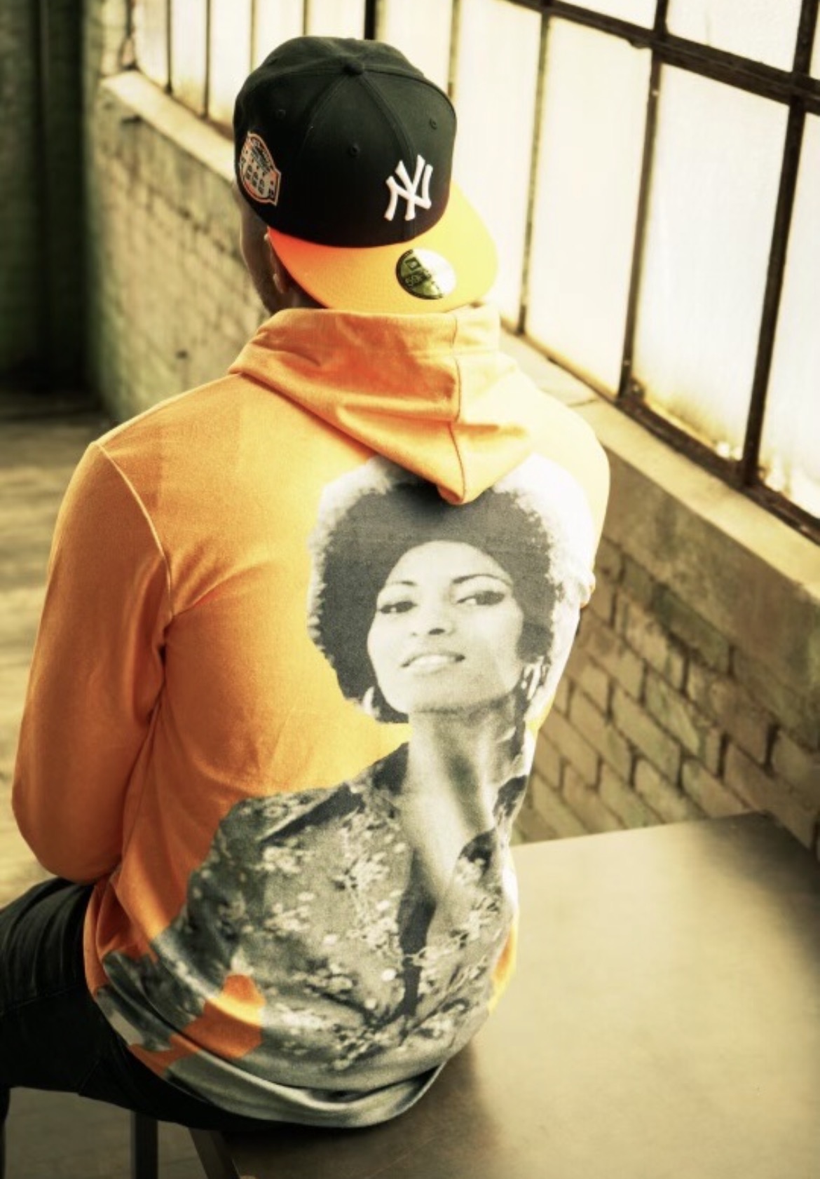 Seated portrait from behind in an orange graphic hoodie and New York cap by a warehouse window