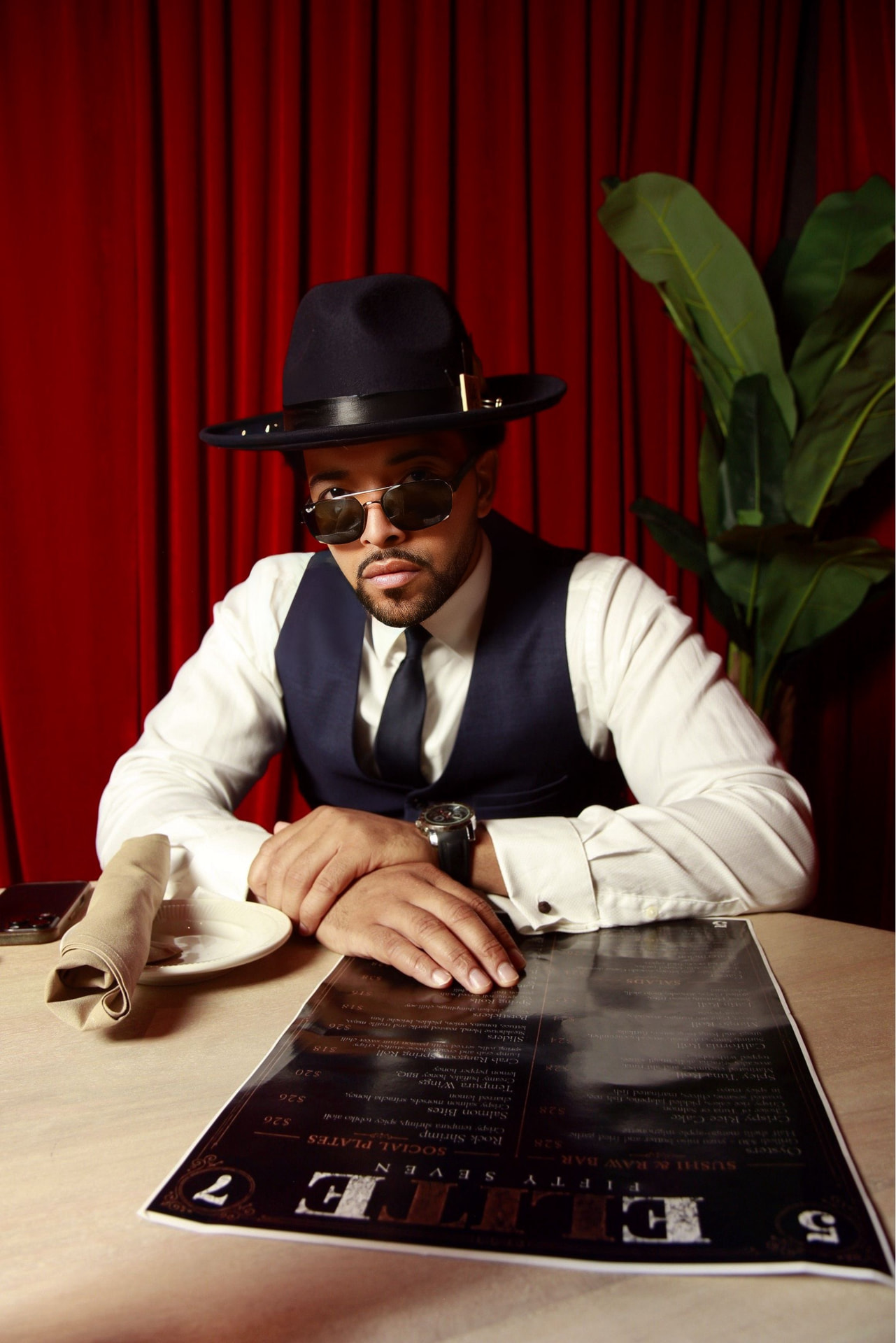 Styled portrait seated at a restaurant table against a red velvet curtain